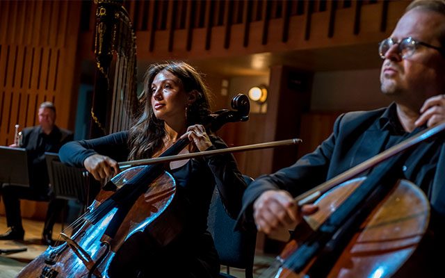 Two cello players sat playing among other musicians, in a wood-panelled hall.