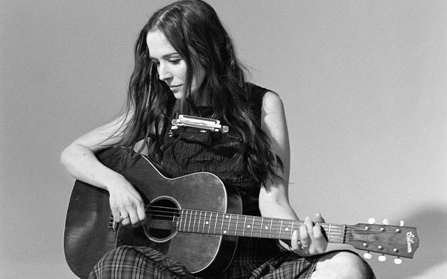 A woman with long dark hair, sitting with her legs folded, holding an acoustic guitar.