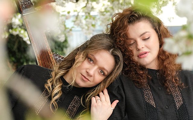 Two women leaning against each other in a leafy outdoor setting, with musical instruments on their backs.