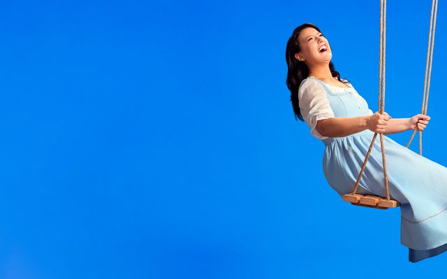 A picture of a woman on a swing smiling. She has long brown hair and is wearing a pale blue dress and brown heeled shoes.