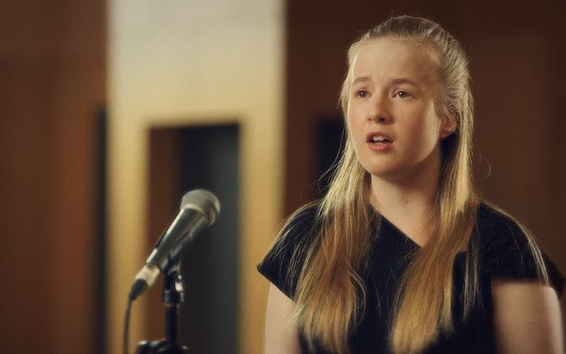 A young woman with long blonde hair wearing a black t-shirt singing into a microphone.