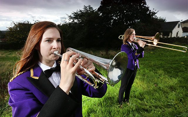 Two women wearing matching band uniforms playing brass instruments in a green outdoor setting.