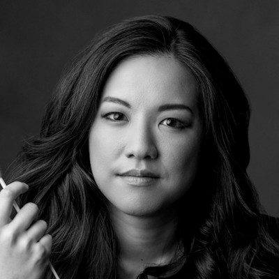 A black and white close-up of a woman with long dark hair, holding a stick-like object out of shot.