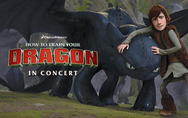 A movie still from How to Train Your Dragon featuring Hiccup, a slender Viking teen in a fur vest, smiling and hugging the head of Toothless, a sleek, black Night Fury dragon. They're standing outdoors on a mossy, green hill with a rocky cliff in the background.