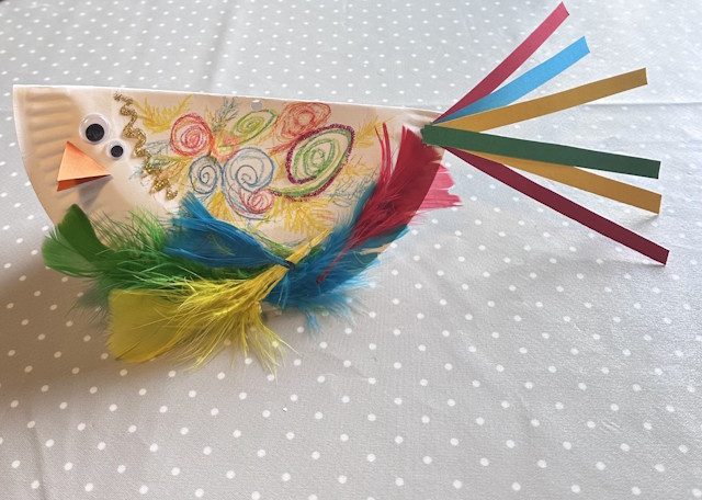 A colourful paper plate bird on a table, with feathers, colourful drawn patterns and paper strips for a tail.