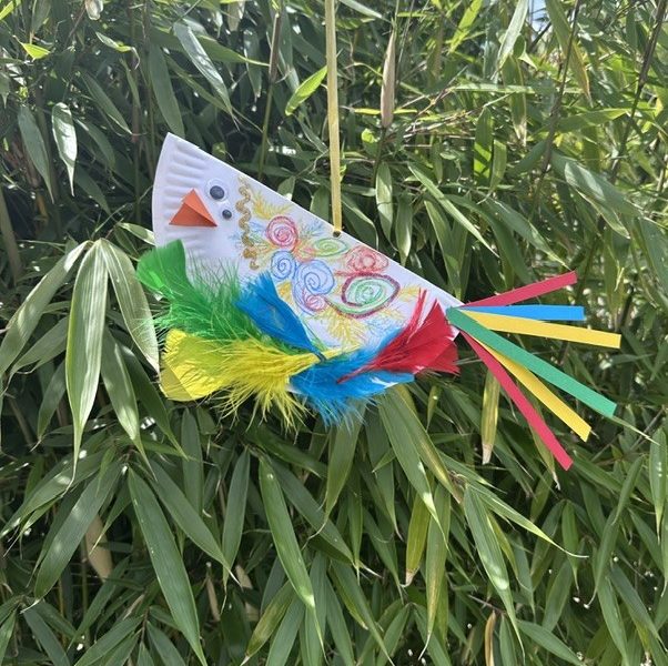 A colourful paper plate bird, in a leafy hedge.