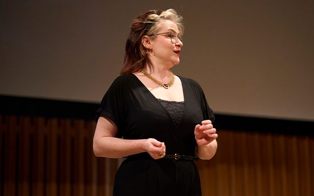 A picture of a woman on a stage wearing a black dress and glasses.
