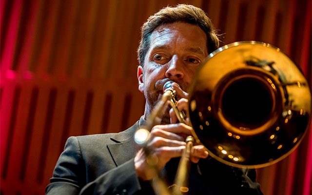 A close-up of a man wearing a dark suit, playing a trombone.