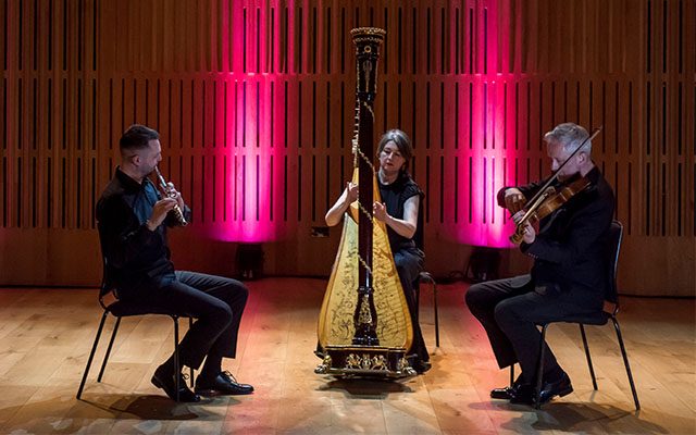 Three musicians playing a flute, harp and violin in front of a backdrop illuminated by red light.