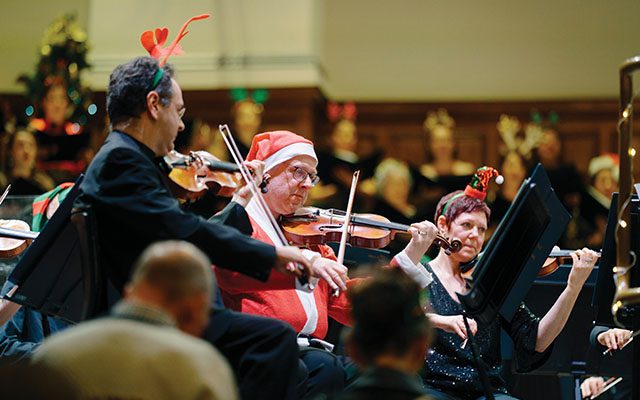 Orchestra members wearing festive Christmas clothing playing inside a large space.