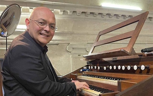 A man wearing a dark shirt sitting at a large organ, smiling.