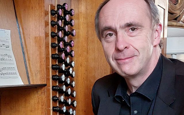 A man wearing a dark suit sat beside a large pipe organ and sheet music.