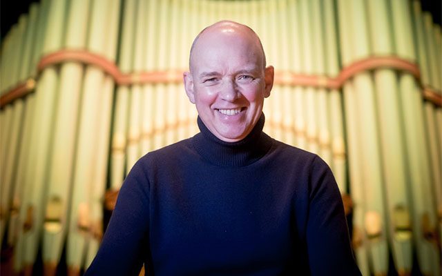 A man wearing a polo-neck jumper standing in front of the pipes of a large pipe organ.