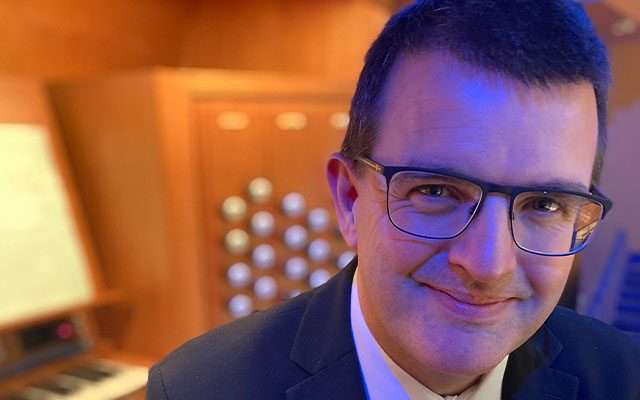 A close-up of a man wearing a dark suit and glasses in front of a pipe organ.