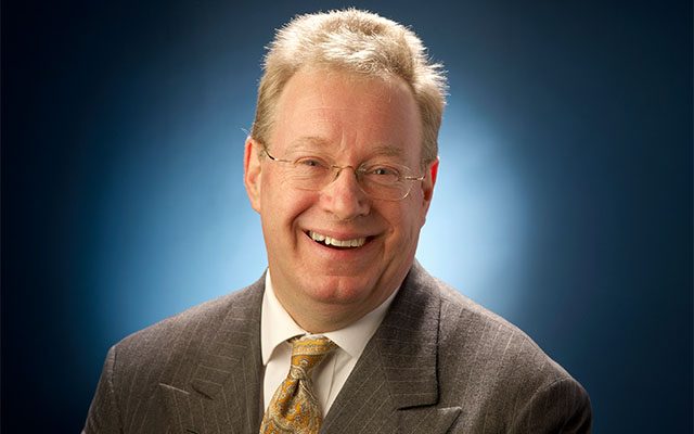 A smiling man with short light-coloured hair and thin-framed glasses, wearing a brown suit and gold tie.