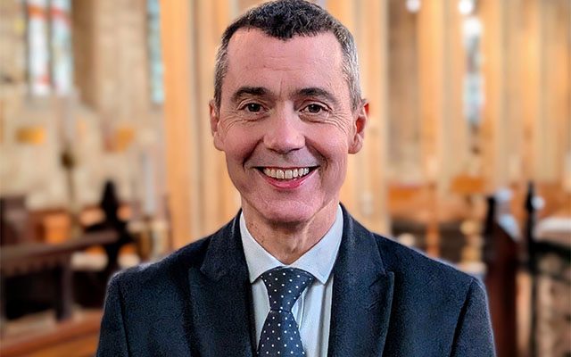 A close-up of a man with short hair wearing a dark suit, smiling inside a church.