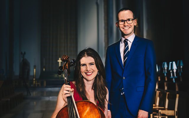 A woman holds a cello beside a man wearing a blue suit and tie inside a hall.