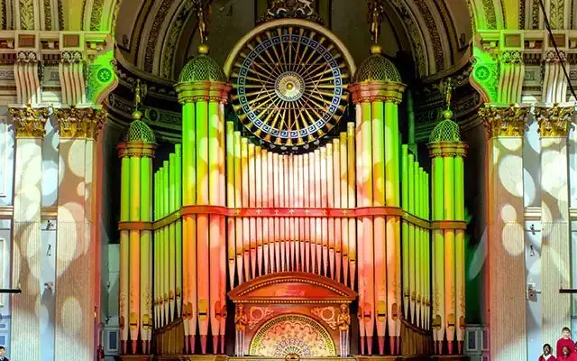 A colourfully illuminated pipe organ inside a town hall, with a large group of young people singing in front of it.