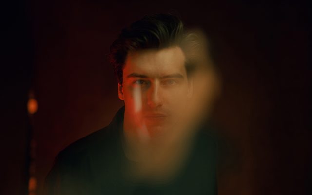 A blurry, stylised image of a man with short dark hair, wearing a dark shirt in front of a dark maroon background.