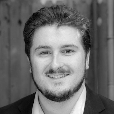 A black and white close-up of a man with short dark hair and a beard, smiling.