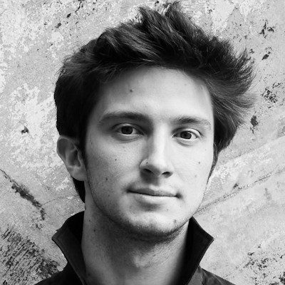 A black and white headshot of a clean-shaven man with short dark hair, wearing a dark shirt.