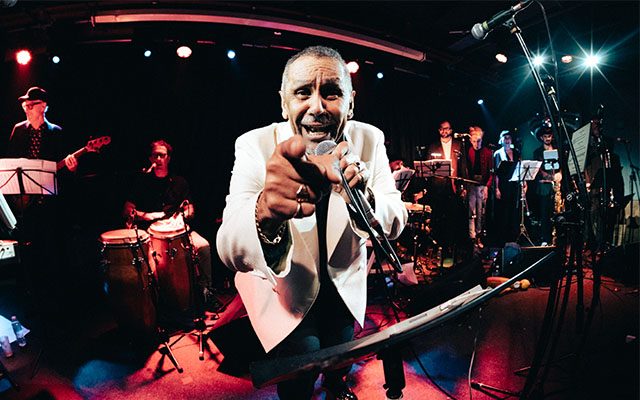 A man on stage wearing a white jacket singing into a microphone in front of a band playing various instruments, including bass guitar, drums and sax.