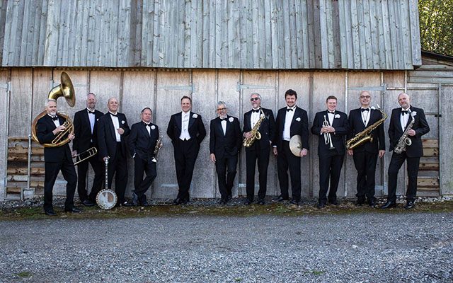 Members of the Pasadena Roof Orchestra in black tuxedos, holding instruments including a banjo, saxophones, and brass, while leaning against a weathered wall.
