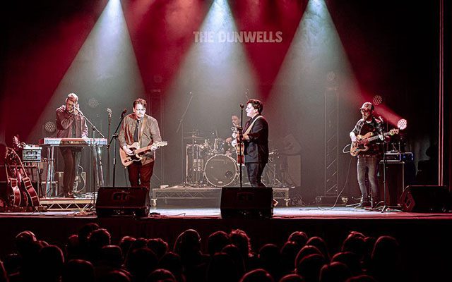 The Dunwells band; a keyboard player, two guitarists, a bassist and drummer performing live on stage in front of a large audience.