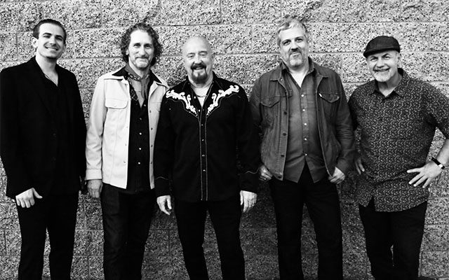 A black and white photograph of The Fabulous Thunderbirds: Five men standing in front of a brick wall, smiling.