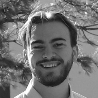 A black and white close-up of a man with short dark hair and a beard wearing a white shirt.