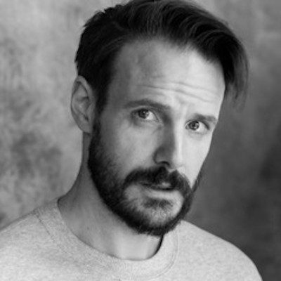 A black and white close-up of a man with short dark hair, a beard and moustache wearing a light-coloured t-shirt.