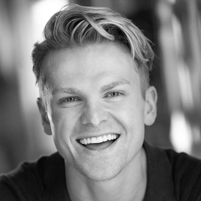 A black and white close-up of a man with medium-length light hair wearing a black t-shirt, smiling.