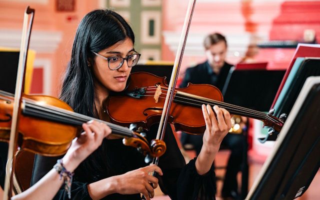 A close-up of a woman playing a violin, surrounded by other orchestral players.