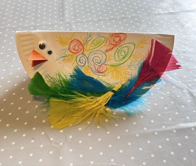 A paper plate with colourful patterns drawn on, feathers, googly eyes and an orange paper beak.