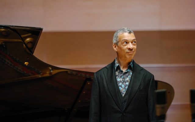 A man with short light hair wearing a dark jacket and blue patterned shirt, standing beside a grand piano.