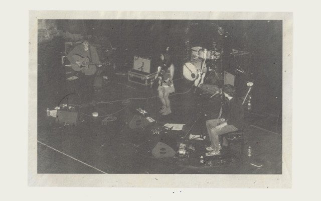 A grainy, black and white photograph of four musicians playing on stage including a drum kit, two guitarists and singer.