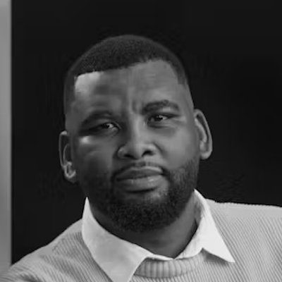 A black and white close-up of a man with short hair and a beard, wearing a white shirt and light-coloured jumper.