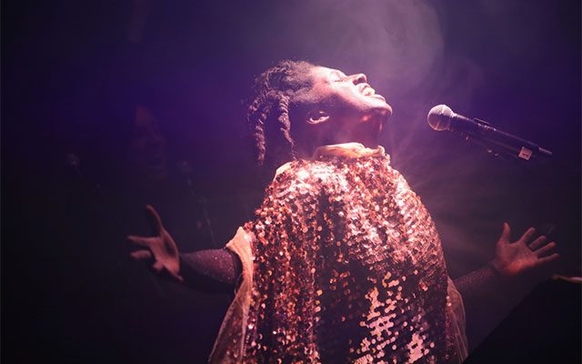 A singer with dreadlocks wearing a shimmering gold sequinned cape, singing passionately with arms outstretched into a microphone.