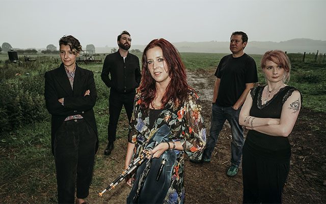 Five individuals standing outside in a flat farmland area with trees.