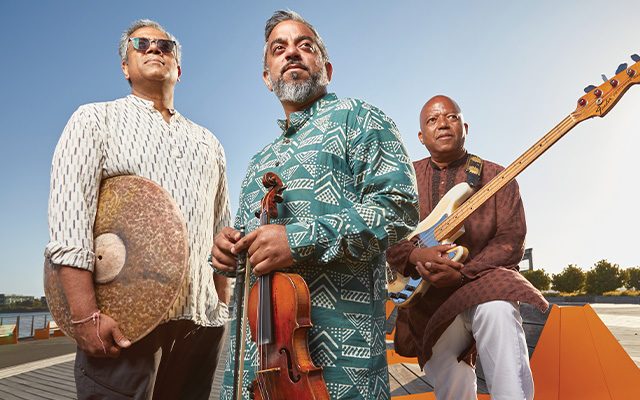 Three men holding instruments - a cymbal, violin and bass guitar, on a pier.