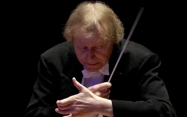 A conductor wearing a black jacket and a white shirt and bow-tie, holding a baton as he conducts.