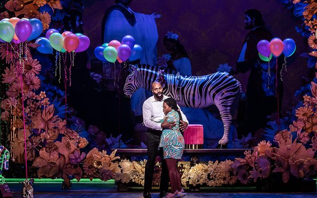 A woman and man embrace on stage in front of a Zebra sculpture and balloons.