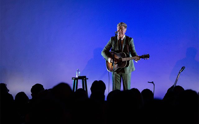 A white man with grey hair and wearing a suit playing guitar and singing.