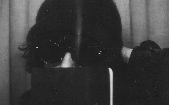 A close up black and white photo of a person wearing a dark beanie and sunglasses with their mouth covered by a notebook